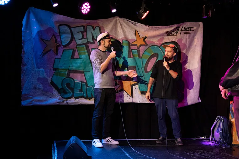 Zwei Rapper mit Mikrofonen vor einem Graffiti-Banner